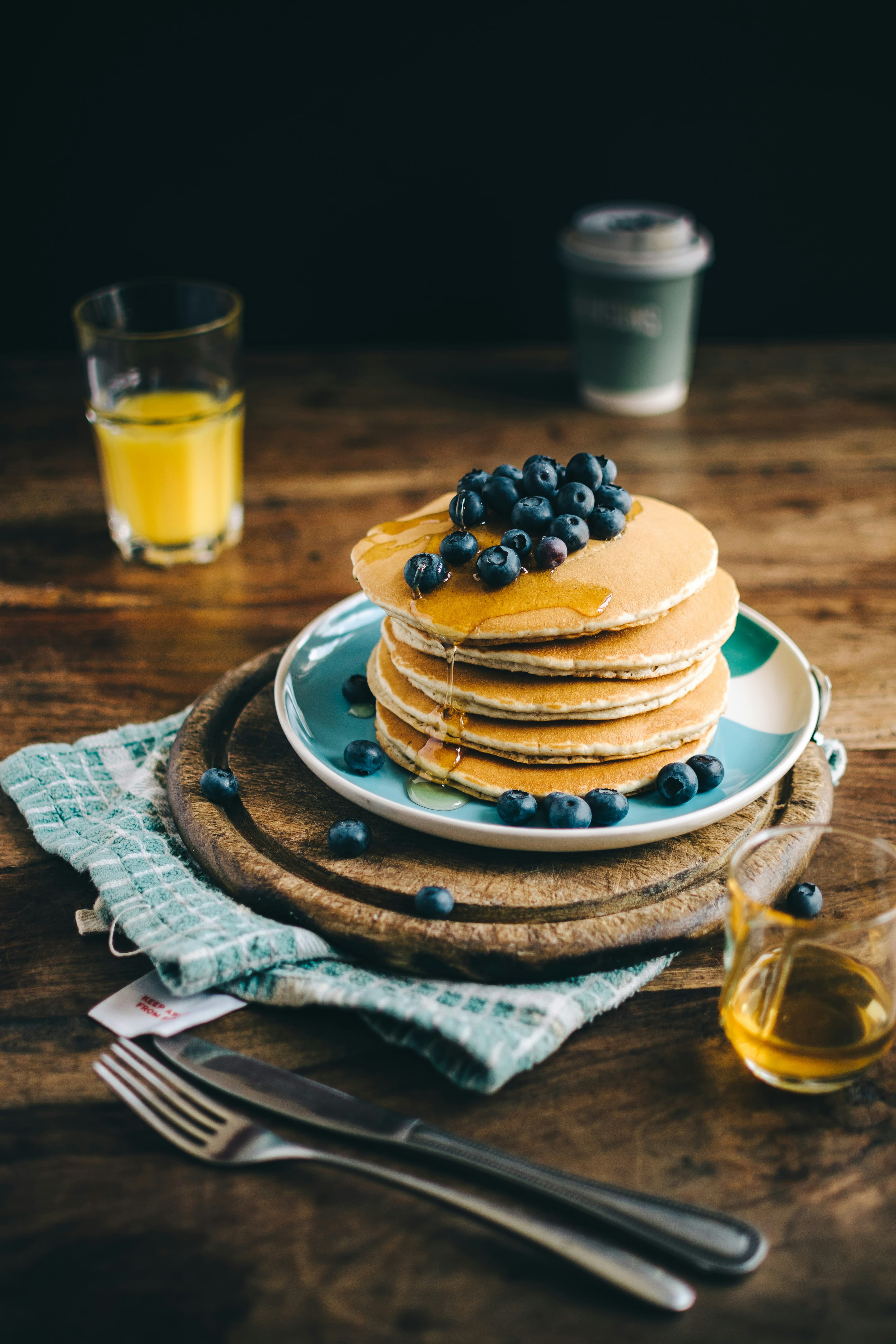 Table en bois dressé avec un plat contenant 5 piles de pancakes dorés, garnis de myrtilles et de miel, une verre de miel.